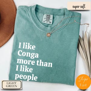 Op de afbeelding: Lichtgroen t-shirt met de tekst "I like Conga more than I like people" in het wit. Het shirt heeft een ronde hals en een klein label op de kraag. Een zilveren ketting met een hanger is over het shirt gedrapeerd.