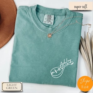 May include: Light green t-shirt with a white mandolin graphic and the name "Ashley". The shirt has a crew neck and short sleeves. A silver necklace with a pendant is draped across the shirt. The words "super soft" are in the upper right corner.