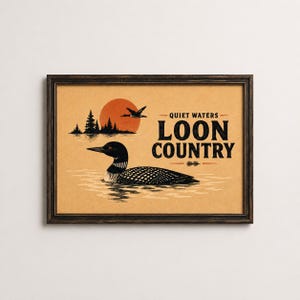 Puede incluir: Una impresión rectangular enmarcada sobre papel color canela que muestra un colimbo negro nadando, un colimbo volando, una silueta de pinos y un gran sol naranja. El texto dice QUIET WATERS LOON COUNTRY en letras negras y negritas.