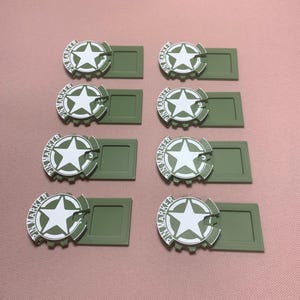 Könnte beinhalten: Acht olivgrüne Pin-Marker mit einem weißen Sterndesign und dem Text "PIN MARKER". Einige Marker haben die Zahlen 5 und 9. Die Marker sind rechteckig mit einem kreisförmigen Design.