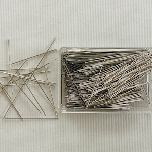 May include: A clear plastic container filled with silver sewing pins. The pins are scattered on a white surface.