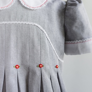 May include: A gray dress with a white collar and white lace trim. The dress has red embroidered flowers and a pleated skirt.