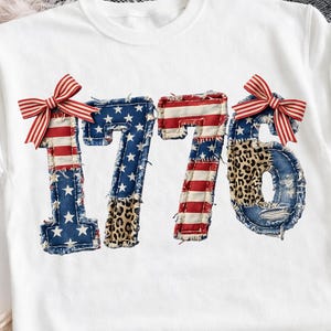 Pode incluir: Camiseta branca com o número "1776" em um design de patchwork. Os números incorporam padrões da bandeira americana, estampa de leopardo e laços listrados em vermelho e branco. Um estilo casual e patriótico.