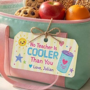 Pode incluir: Uma lancheira isolada turquesa e rosa cheia de biscoitos, uma maçã e uma laranja. Dentro, uma garrafa de água com uma fatia de limão. Uma etiqueta de presente diz "No Teacher Is COOLER Than You" com um desenho de uma bebida. Lápis e um vaso de tulipas cor-de-rosa estão por perto.