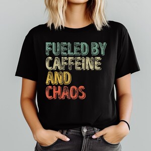 May include: Black t-shirt with the text "FUELED BY CAFFEINE AND CHAOS" in a retro, multi-colored design. The shirt is a crew neck and is being worn by a person with blonde hair.