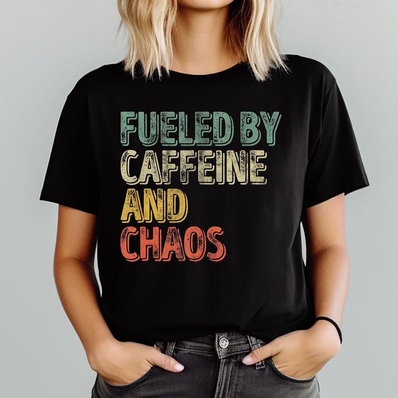 May include: Black t-shirt with the text "FUELED BY CAFFEINE AND CHAOS" in a retro, multi-colored design. The shirt is a crew neck and is being worn by a person with blonde hair.
