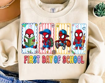 May include: Cream-colored sweatshirt featuring a colorful graphic of Spider-Man characters in school-themed poses. The phrase "FIRST DAY OF SCHOOL" is printed in a rainbow font across the bottom of the design.