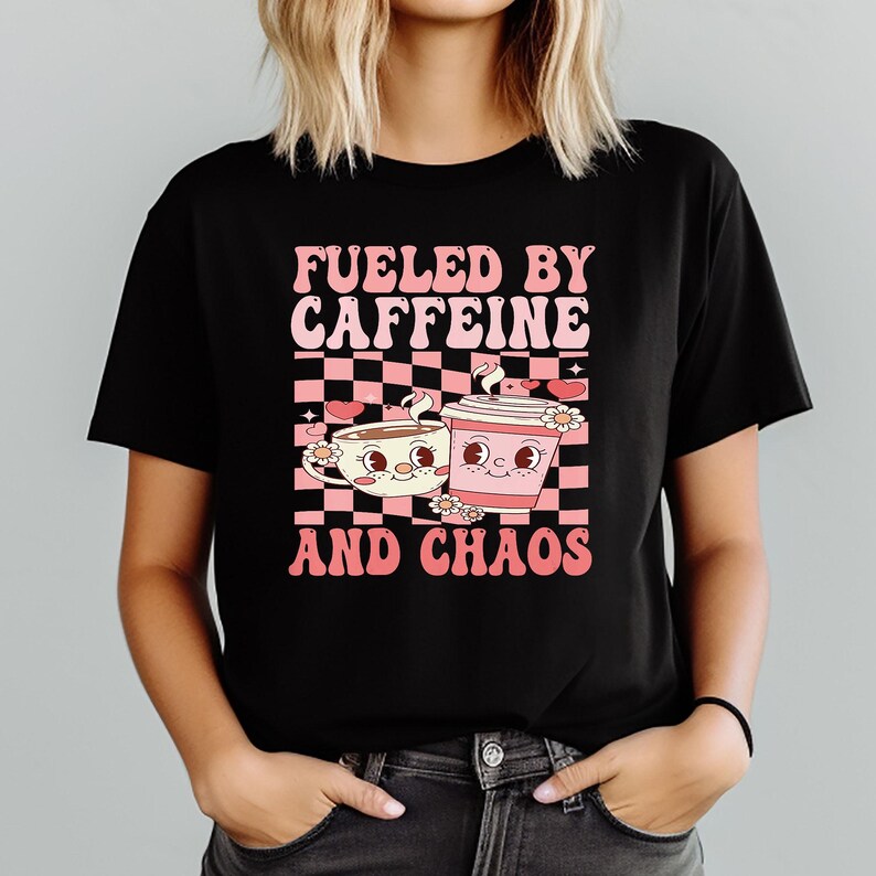 May include: Black t-shirt with a retro design featuring two cartoon coffee cups and the text "FUELED BY CAFFEINE AND CHAOS" in pink and white. The design includes a checkered background and floral accents.