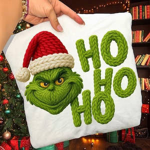 Pode incluir: Almofada quadrada branca com um personagem verde do Grinch usando um chapéu de Papai Noel vermelho com uma borda branca. A almofada também tem as palavras "HO HO HO" em letras verdes em estilo de malha. A almofada é segurada por uma mão.