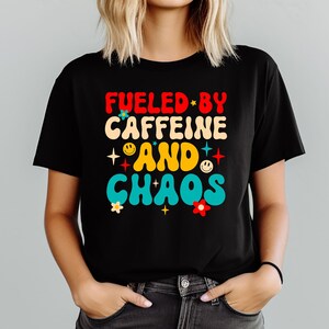 May include: Black t-shirt with the text "FUELED BY CAFFEINE AND CHAOS" in red, cream, and turquoise lettering. The design includes stars, smiley faces, and flowers. A casual, everyday tee.