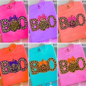 Pode incluir: Camisolas de gola redonda em várias cores, cada uma com a palavra "BOO" com um design de estampa de leopardo. O "O" é uma abóbora ou fantasma com um laço. As camisolas estão disponíveis em rosa, roxo, laranja e azul-petróleo.