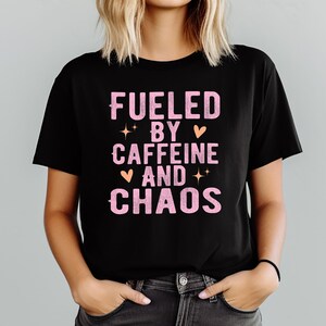 May include: Black t-shirt with the text "FUELED BY CAFFEINE AND CHAOS" in pink, with star and heart graphics. The shirt is a crew neck and is being worn.