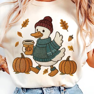 Pode incluir: Camiseta creme com um pato bordado usando um gorro bordô, cachecol e suéter verde-azulado, segurando uma xícara de café. Duas abóboras e folhas de outono bordadas, criando um tema de outono.