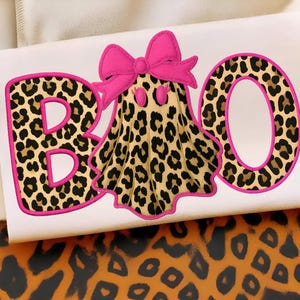 Pode incluir: Um design com tema de Halloween com a palavra "BOO" em estampa de leopardo e contornos rosa. Um fantasma com um lençol de estampa de leopardo e um laço rosa está no centro. O design está em um fundo branco.