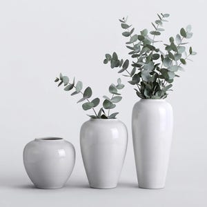 Op de afbeelding: Drie gladde, glanzend witte keramische vazen van verschillende hoogtes op een rij. De twee langste vazen bevatten takjes met groene eucalyptusbladeren met afgerond blad, terwijl de kleinste vaas aan de linkerkant leeg blijft.