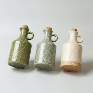 May include: Three ceramic cruets with handles and cork stoppers. The first is a glossy dark green, the second a textured sage green, and the third a cream-coloured vessel with an exposed terracotta base. Each features a narrow neck and spout.