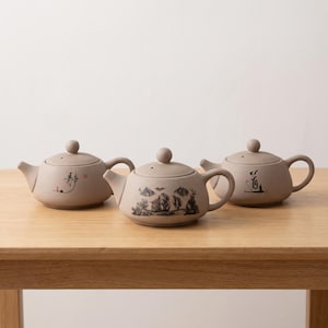 May include: Three matte light brown ceramic teapots with lids and curved handles. The left teapot features black calligraphy and a small red stamp. The centre teapot displays a detailed black landscape illustration. The right teapot has black calligraphy.
