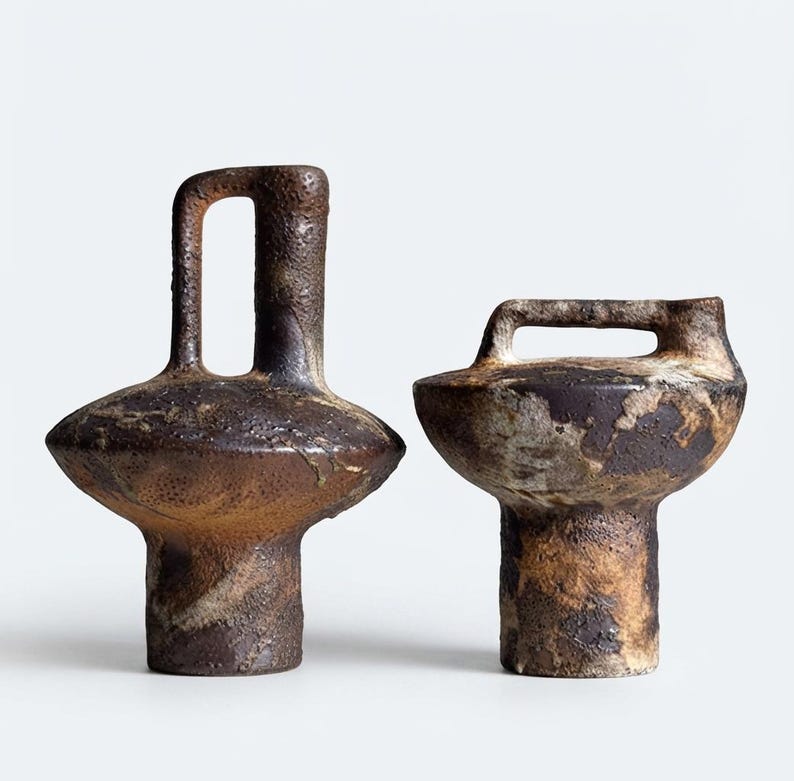 May include: Two ceramic vessels with a rough, textured surface in shades of dark brown, tan, and cream. The taller vessel features a vertical handle, while the shorter, wider vessel has a horizontal handle across its top opening.