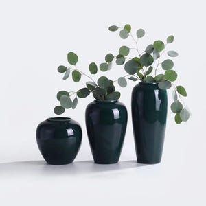 May include: Three dark green ceramic vases of varying heights and shapes. The shortest vase is round, the middle is oval, and the tallest is cylindrical. Each vase contains green eucalyptus branches with round leaves, creating a cohesive set.