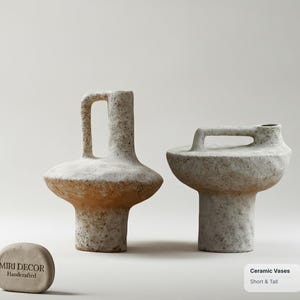 Könnte beinhalten: Zwei Keramikvasen mit einer strukturierten, gesprenkelten cremeweißen Oberfläche. Eine Vase ist hoch mit einem vertikalen Henkel, die andere ist kurz mit einem horizontalen Henkel. Ein kleiner steinartiger Gegenstand trägt den Text MIRI DECOR Handcrafted.