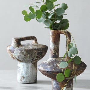 May include: Two ceramic vessels with a textured, mottled brown and white glaze. Each piece features a wide, rounded midsection, a cylindrical base, and a vertical handle extending from the top. A sprig of green eucalyptus leaves rests inside.