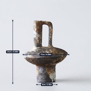 May include: A textured ceramic vase with a dark brown and mottled grey finish. It features a central handle and a wide, disc-shaped body. The vase measures 9.06 inches in height, 6.69 inches in width, and has a base diameter of 3.15 inches.