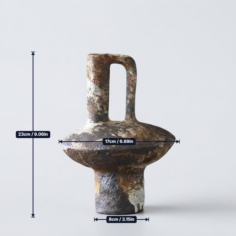 May include: A textured ceramic vase with a dark brown and mottled grey finish. It features a central handle and a wide, disc-shaped body. The vase measures 9.06 inches in height, 6.69 inches in width, and has a base diameter of 3.15 inches.