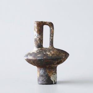 May include: A ceramic vessel with a textured, dark brown and mottled white finish. The object features a cylindrical base, a wide, flattened midsection, and a tall, narrow neck with a single vertical handle attached to the side of the neck.