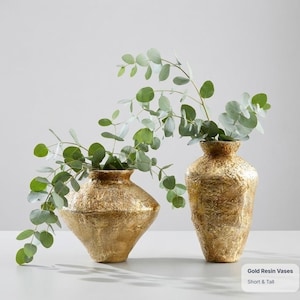 Op de afbeelding: Twee gouden harsvazen met een getextureerde, gehamerde afwerking. De ene vaas is kort en breed met een taps toelopende voet, terwijl de andere hoog en slank is. Beide vazen bevatten groene eucalyptus takken. De tekst luidt Gold Resin Vases.