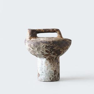 May include: A small, hand-formed ceramic vessel with a textured, mottled brown and grey surface. The object features a cylindrical base that widens into a rounded, bowl-like top section, finished with a single arched handle on one side.