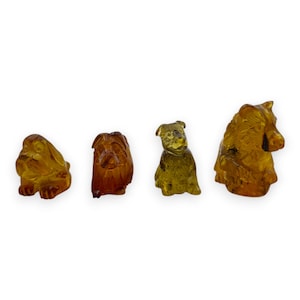May include: Four amber-colored dog figurines in various poses. The dogs are carved with detail, showcasing different breeds. The amber has a translucent quality, allowing light to pass through, highlighting the intricate carvings.