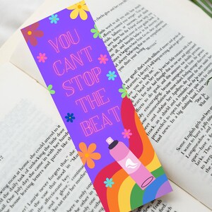 May include: A colourful bookmark featuring the phrase "YOU CAN'T STOP THE BEAT". It has a purple background with a rainbow, flowers, and a pink spray bottle illustration. Ideal for marking pages in a book.