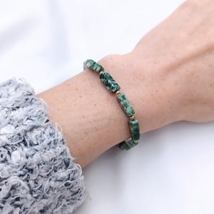 Könnte beinhalten: Ein grün-weißes Perlenarmband mit goldenen Akzenten. Das Armband hat rechteckige Perlen mit einem gesprenkelten Muster. Es wird am Handgelenk getragen. Ein grauer Ärmel ist im Hintergrund zu sehen.
