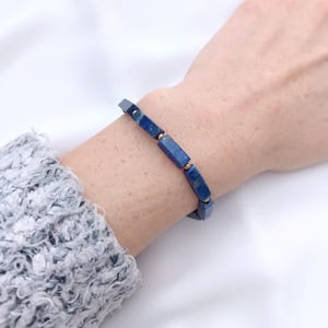 Può includere: Un braccialetto in lapislazzuli blu con perline rettangolari e piccoli distanziatori color oro. Il braccialetto è indossato su un polso, mettendo in risalto il colore e il design del gioiello. Il braccialetto è un gioiello di moda.