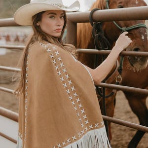 May include: A tan suede poncho with white fringe and decorative stitching. The poncho is worn with a white cowboy hat. A brown horse is in the background, near a metal fence. The overall theme is Western wear.