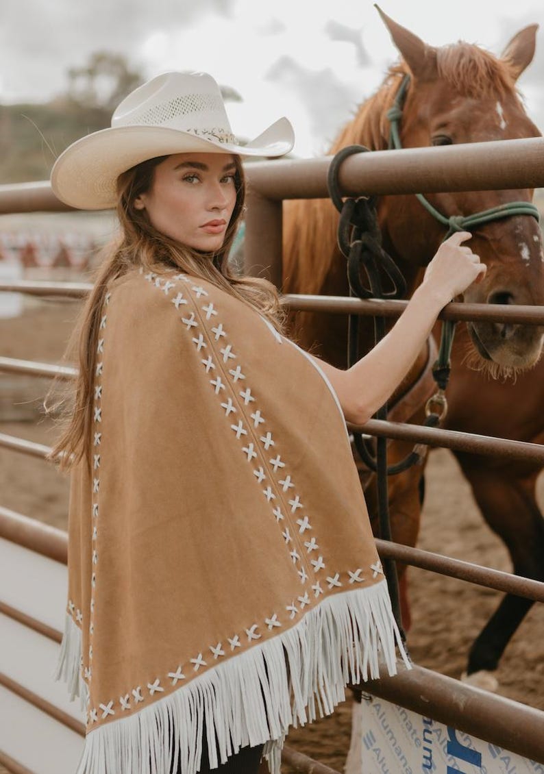 May include: A tan suede poncho with white fringe and decorative stitching. The poncho is worn with a white cowboy hat. A brown horse is in the background, near a metal fence. The overall theme is Western wear.