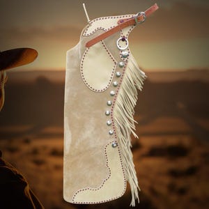 May include: A tan suede leather chaps with cream-colored accents and decorative stitching. The chaps feature silver-colored studs, a fringe along the side, and a brown leather belt with a buckle. The background is a blurred landscape.