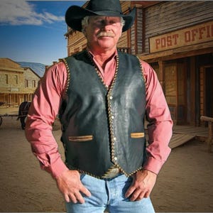 Peut inclure: Un homme portant un gilet en cuir noir avec des surpiqûres brunes et une chemise boutonnée rose clair. Le gilet a une bordure tressée et deux poches avec des rabats en cuir brun. L'homme porte un chapeau de cowboy et un jean bleu. L'arrière-plan est une ville à thème western.