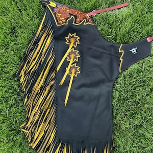 Peut inclure: Chaps en cuir noir avec de longues franges jaunes et noires sur le côté. La partie supérieure présente un motif floral marron gravé avec une bordure jaune. Trois appliques en forme d'étoile avec des accents jaunes ornent le panneau avant.