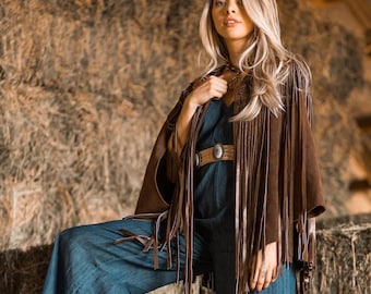 May include: A brown suede fringed cape with a matching belt and a blue jumpsuit. The cape has long fringe detailing along the edges. The belt has a large oval buckle. The jumpsuit has wide legs.