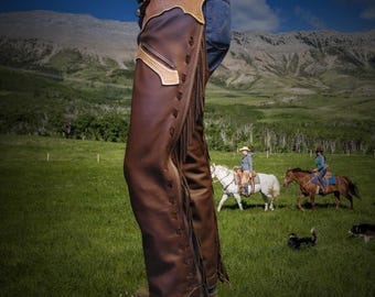 May include: Brown leather chaps with fringe detail. The chaps are worn over blue jeans and a belt with a decorative buckle. The background shows a green field, mountains, and people riding horses.