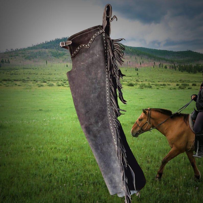 May include: A pair of brown leather chaps with white stitching and fringe. The chaps are hanging in a field with a horse and rider in the background.