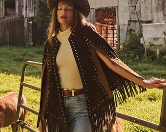 May include: A brown suede cape with fringe and stud details. The cape is worn over a cream-colored turtleneck and paired with blue jeans and a brown belt. A brown cowboy hat completes the look.