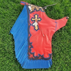 Pode incluir: Um par de perneiras de couro com um painel principal vermelho vibrante, uma grande aplicação de cruz bege e um painel lateral azul com franjas longas. O design inclui acabamento preto recortado, detalhes florais e uma tira com fivela.