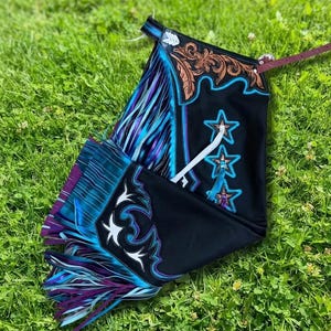May include: Black leather chaps featuring a brown floral tooled leather yoke, turquoise leather star appliqués, and long fringe in shades of blue and purple. The garment includes a thin maroon adjustable strap with a silver-toned metal buckle.