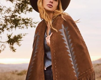 May include: A brown suede cape with fringe and decorative stitching. The cape features dark gray accents and a unique cut-out design. A brown hat completes the western-inspired look.