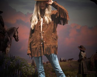 May include: A brown suede jacket with fringe detailing. The jacket has a button closure and a pointed collar. The model is wearing a white cowboy hat and blue jeans.