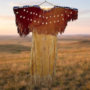 Könnte beinhalten: Ein beiges Wildlederkleid mit einer dunkelbraunen Passe, weißen Muschelverzierungen und langen Fransen an Ärmeln und Saum. Das Kleidungsstück hat eine blau-weiße Perlenborte am Ausschnitt und vertikale Perlenstränge im unteren Bereich.