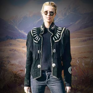 Puede incluir: Una chaqueta de ante negro con detalles de flecos y acentos de cuentas blancas. La chaqueta tiene una cremallera y dos bolsillos. La modelo lleva la chaqueta con un suéter negro y jeans azules.