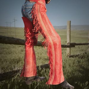 May include: Red leather chaps with fringe detailing and a decorative studded border. The chaps are worn over blue jeans and black boots. The image is set against a backdrop of a green field and a blue sky.